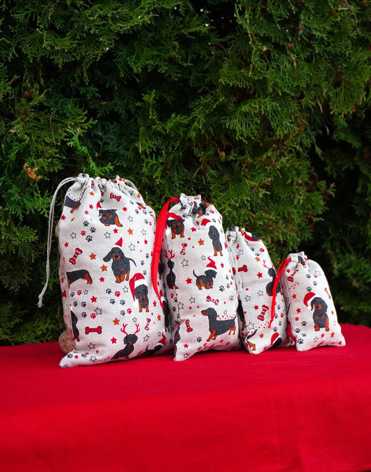 Four drawstring gift bags in descending sizes, printed with black dachshund dogs wearing red Santa hats and surrounded by red stars and bones on a white background, set against a red fabric surface and a green pine background.