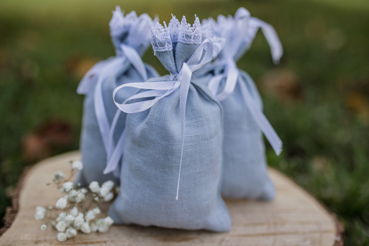 Three small dusty blue linen drawstring gift bags with lace trim and white ribbon bows, arranged on a rustic wooden slice outdoors with blurred green grass in the background.