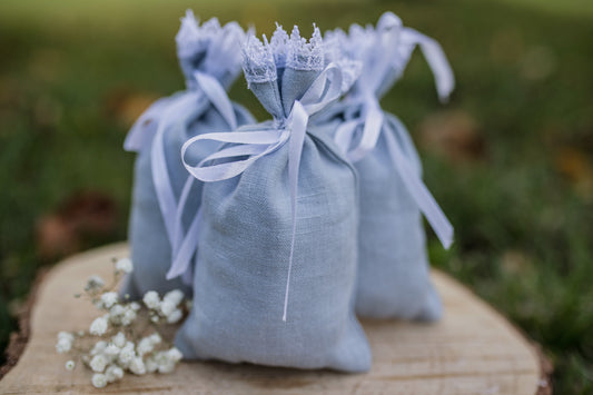 Three small dusty blue linen drawstring gift bags with lace trim and white ribbon bows, arranged on a rustic wooden slice outdoors with blurred green grass in the background.
