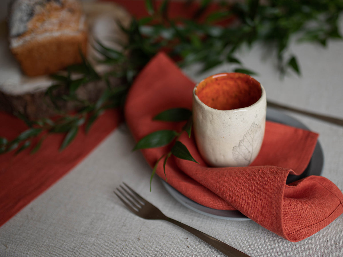 Table setting with a white ceramic cup on a clay linen  napkin, surrounded by greenery.
