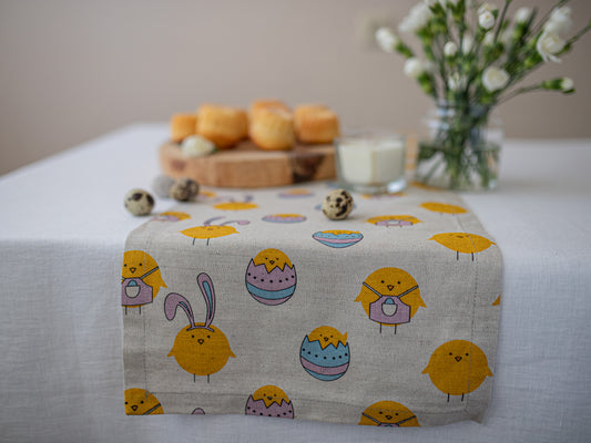 A light beige linen table runner with a cute Easter print of yellow chicks, bunnies, and colorful eggs, styled on a white tablecloth with bread, quail eggs, and flowers in the background.