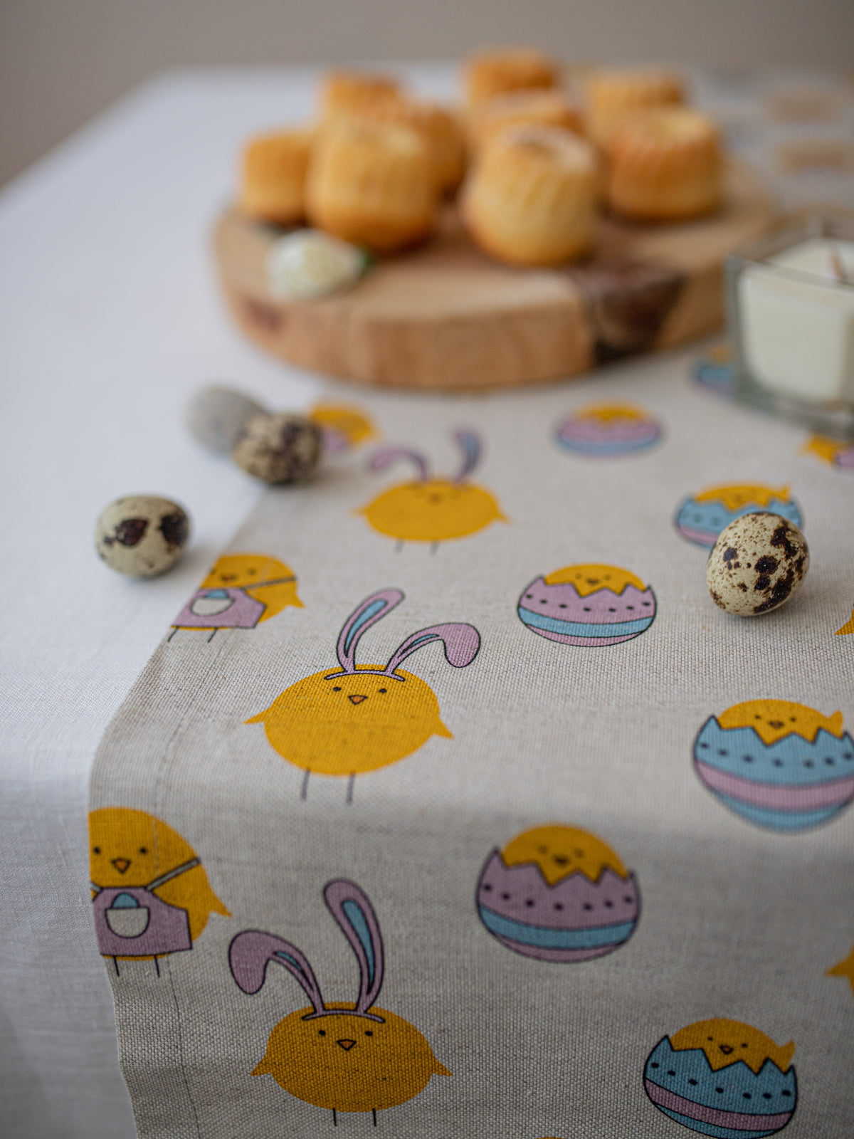 A light beige linen table runner with a colorful Easter print featuring cartoon bunnies, chicks, and decorated eggs, styled on a dining table with quail eggs and a wooden board in the background.