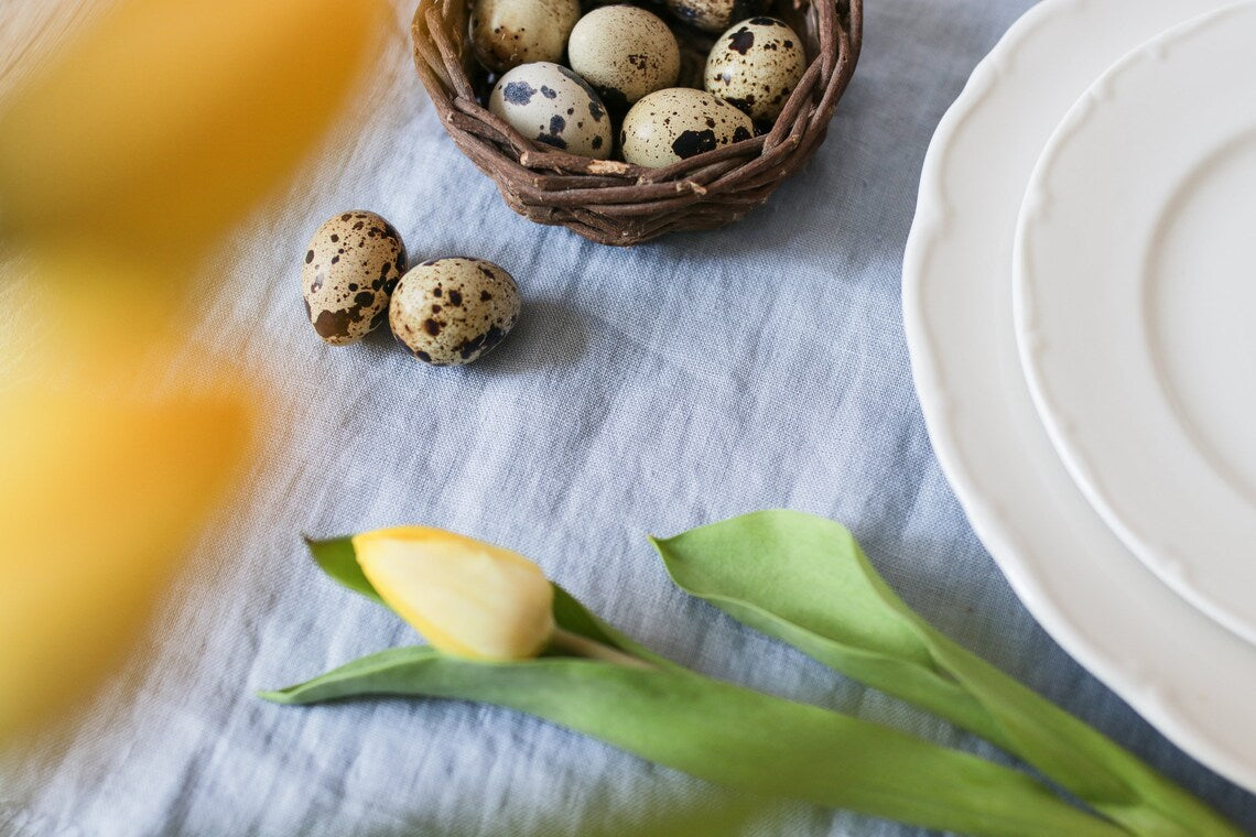 A styled dusty blue table runner scene with speckled quail eggs in a small woven basket, a few loose eggs beside it, white plates on the right, tulip flowers in the foreground, and a light blue fabric surface underneath.