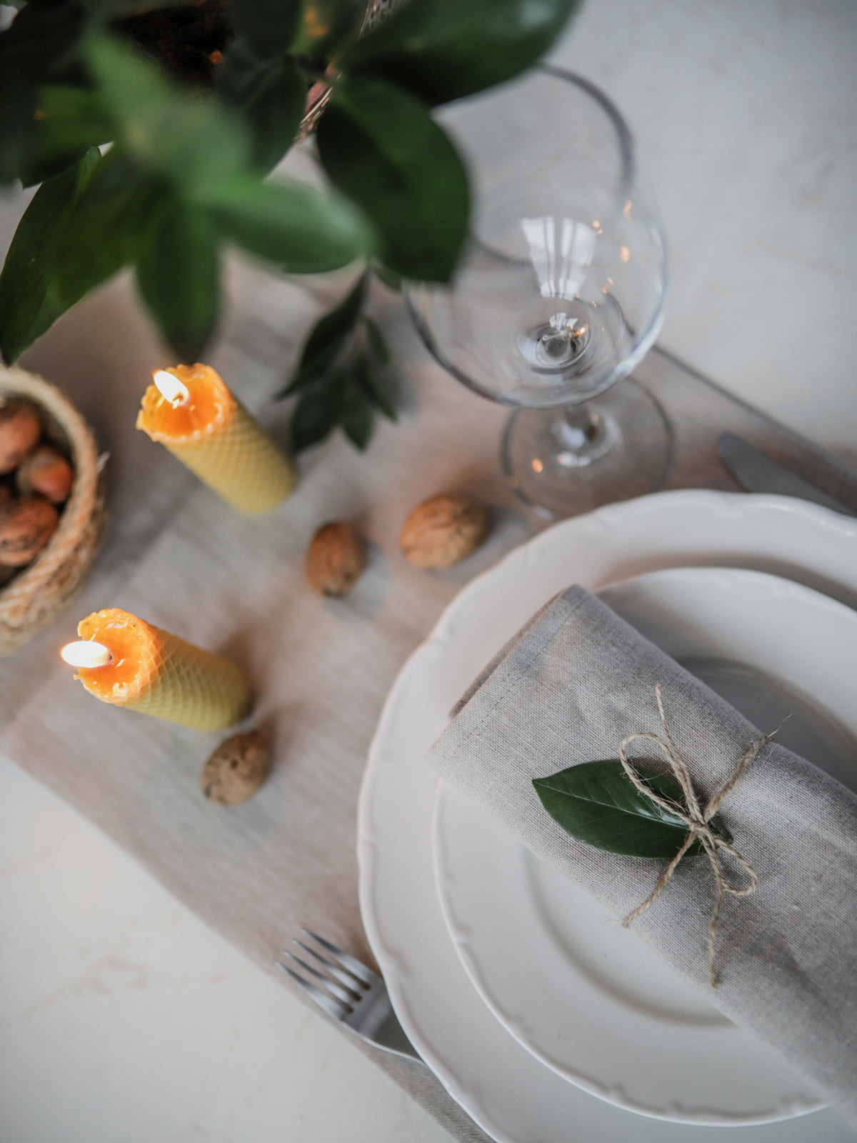 Table setting with white plates, not dyed linen napkin, a glass, and decorative candles on a light surface.