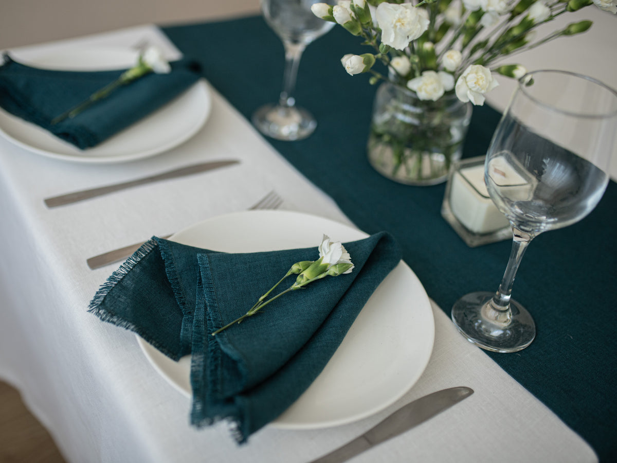 A styled dining table setting with white plates, dark teal/emerald green linen  napkins with fringe, silver flatware, a white floral centerpiece in a glass jar, and clear wine glasses on a white tablecloth with a dark teal runner.