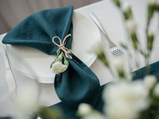 Teal/emerald green linen napkin with a decorative bow and white flowers on a white plate with a fork, blurred floral background.
