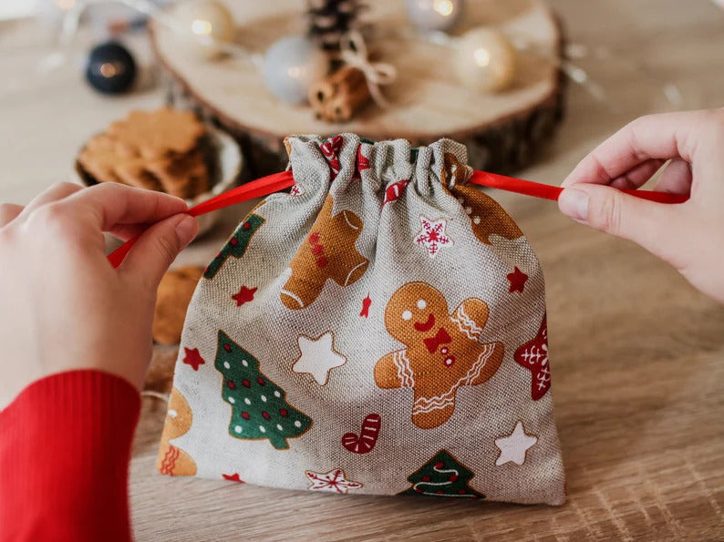 A small linen fabric drawstring bag with a Christmas gingerbread man and tree print, held open by two hands against a warm wooden holiday background.