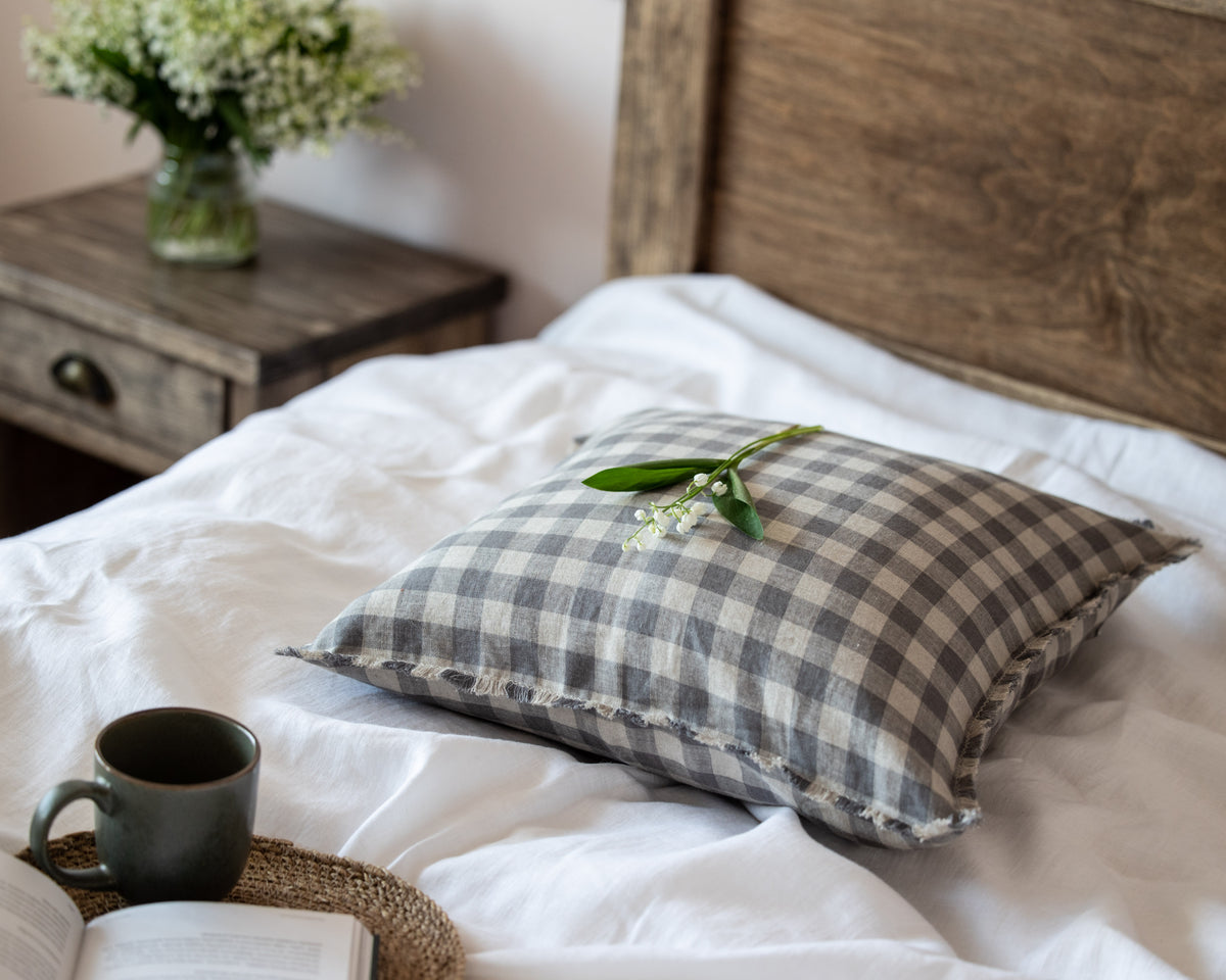 Checkered  linen pillow on a bed with a mug and book in a cozy bedroom setting.