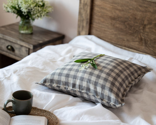 Checkered  linen pillow on a bed with a mug and book in a cozy bedroom setting.