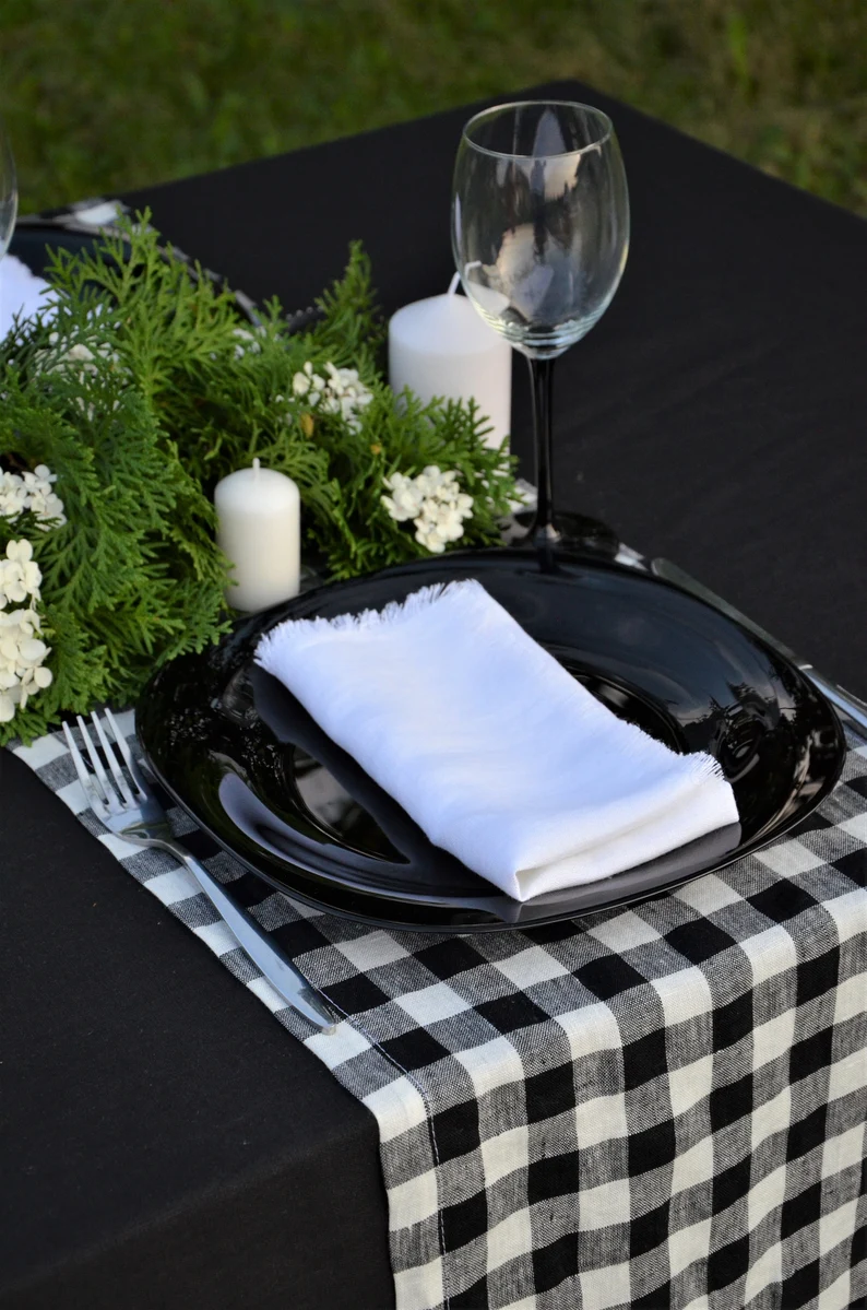 A styled table setting featuring a black oval charger plate with a folded white napkin, a black-and-white checkered runner, white candles, green foliage, and a wine glass on a black tablecloth.