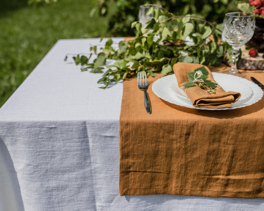 Table setting with a brown linen tablecloth, white plate, mustard linen napkin, and greenery.