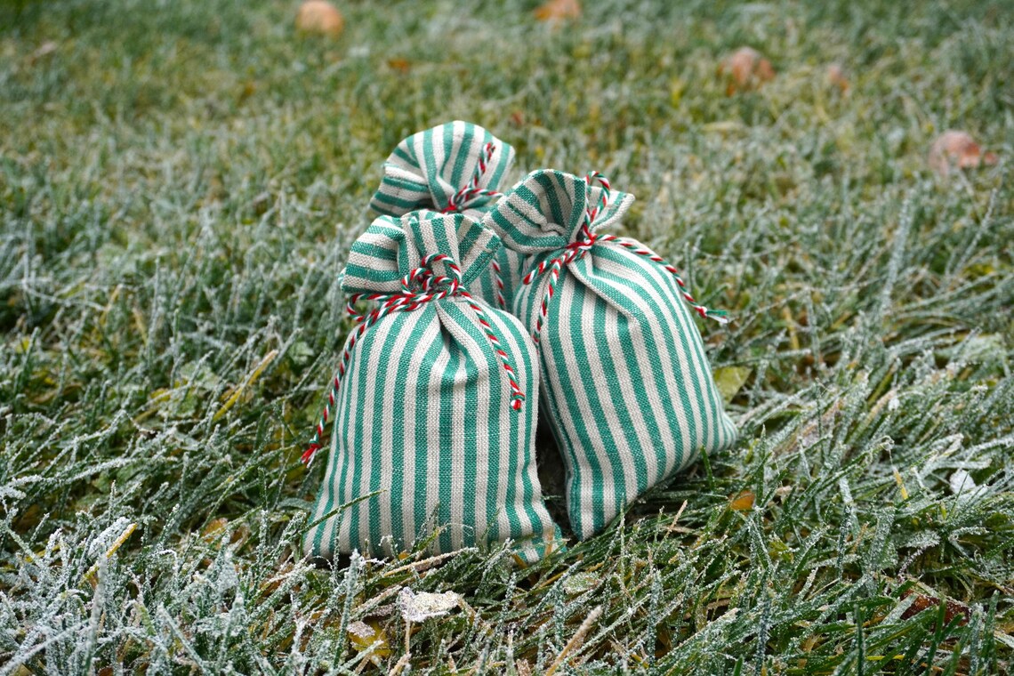 Three small linen fabric drawstring bags with green and white vertical stripes sit on frosty grass outdoors.