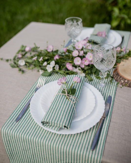 A styled outdoor table setting featuring a green and white striped table runner and matching napkin tied with twine, white plates, clear cut-glass wine glasses, and a floral centerpiece with pink blossoms and greenery.