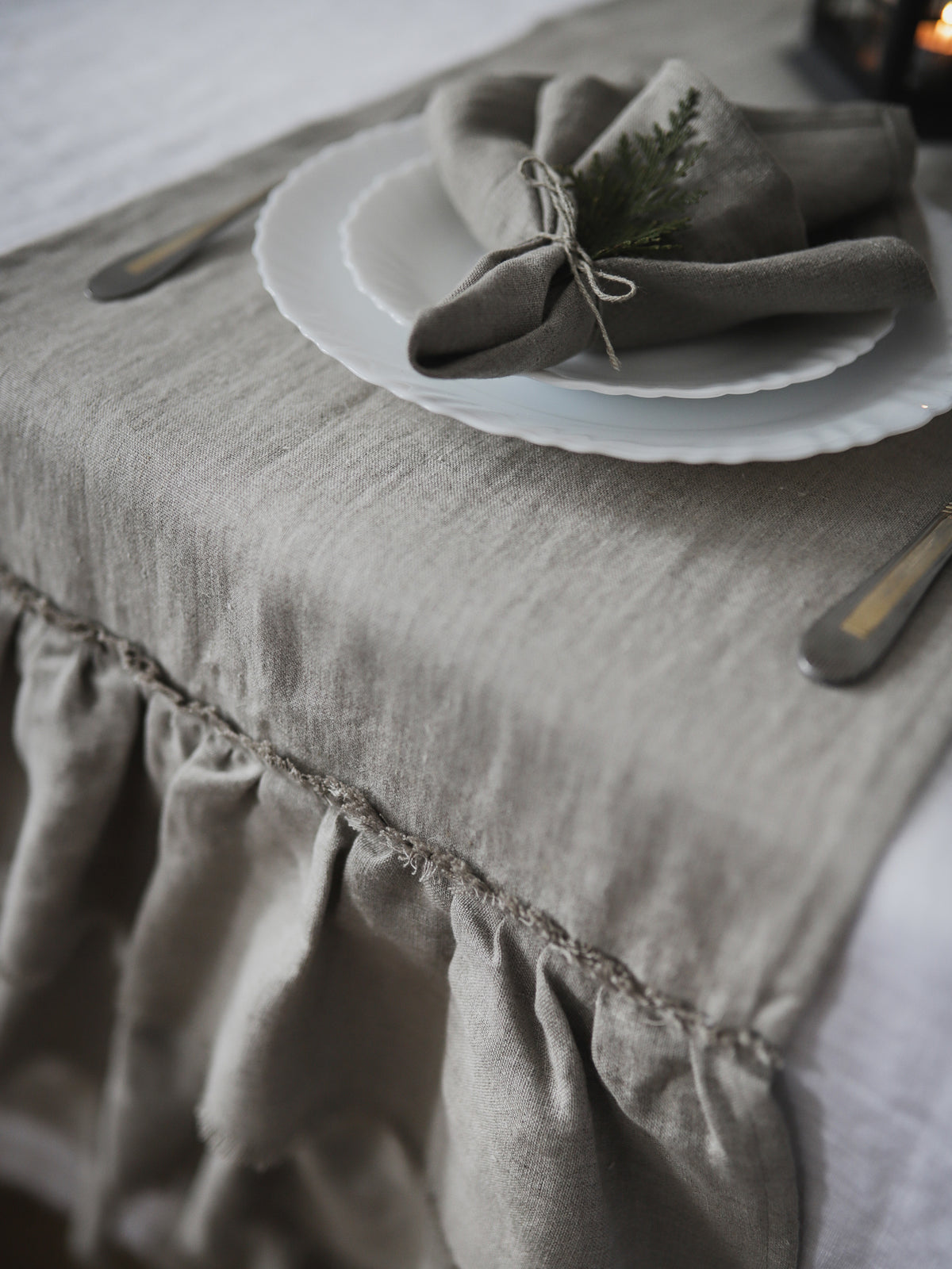 A close-up of a natural color  linen table runner with a ruffled hem, styled on a white tablecloth with a white plate, a folded taupe linen napkin tied with twine, and a small green sprig.