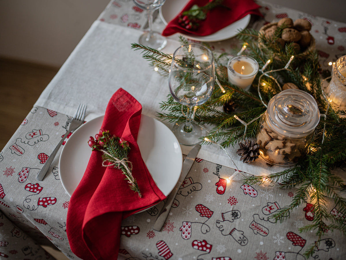 A festive Christmas table setting featuring a white plate with a red linen napkin, decorated with a small evergreen sprig and red berries. The table is styled with a patterned tablecloth, pine branches, warm string lights, and glass jars.