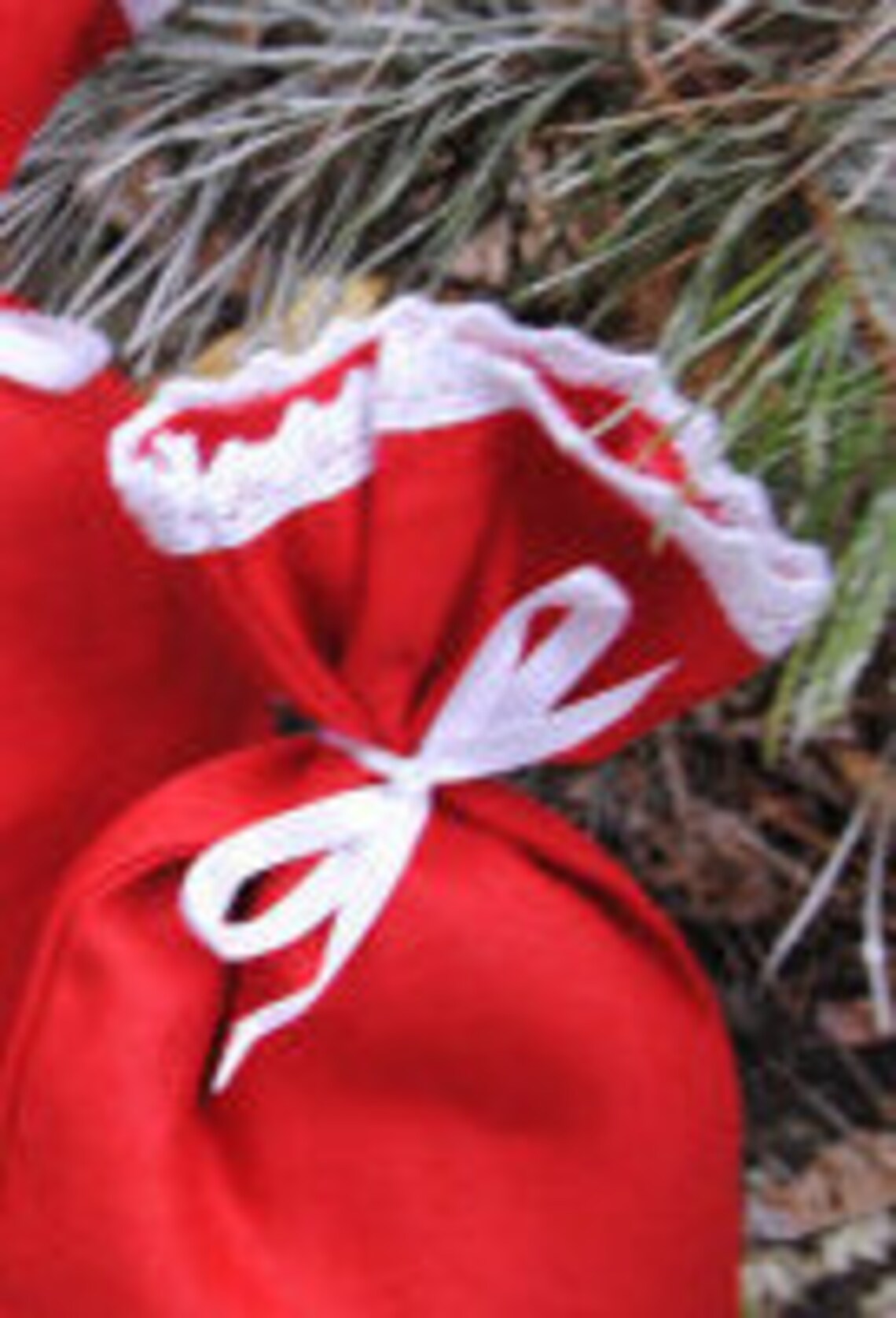 A close-up of a red lien fabric Cristmas pouch with white lace trim and a white ribbon bow, photographed outdoors against pine branches and leaves.