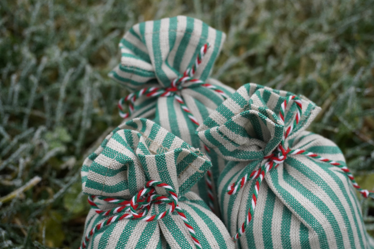 Three small linen drawstring pouches with green and white stripes tied with red and white bakers twine, resting on a frosty outdoor grassy surface.