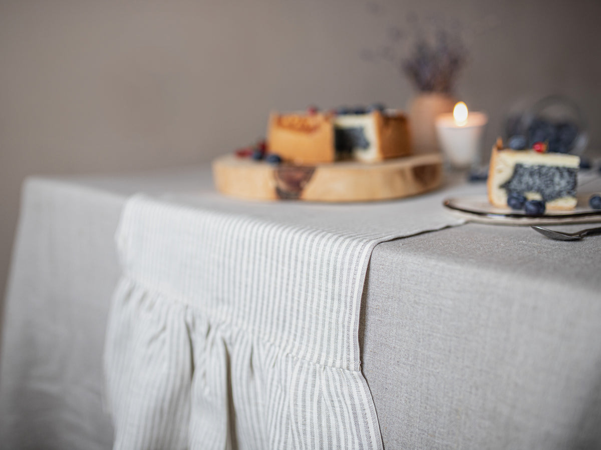 A stripes linen table runner, styled on a table with a wooden board holding a slice of cheesecake and a candle in the background.