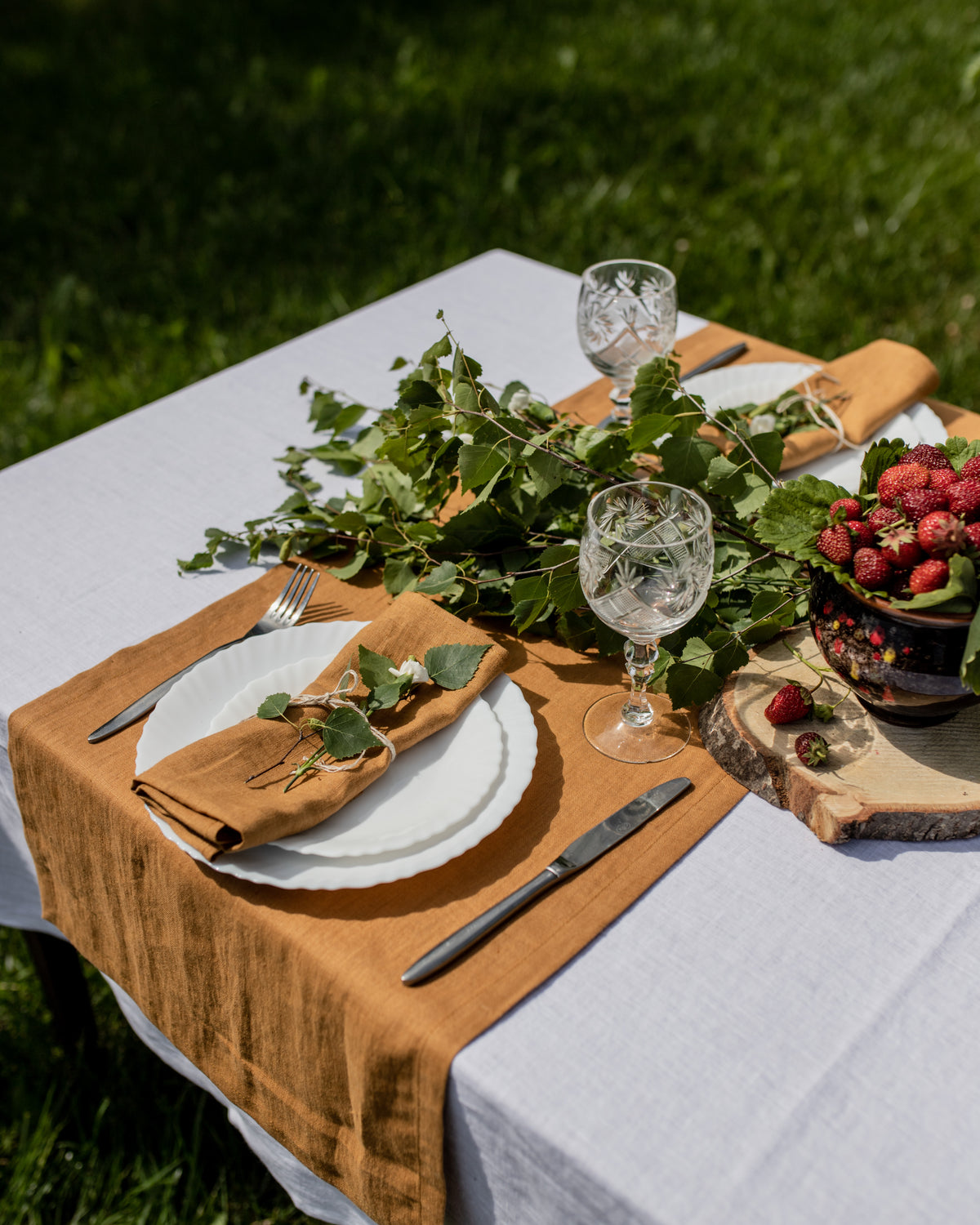 Outdoor table setting with greenery, mustard linen  napkin, white plates, and glasses on a grassy background