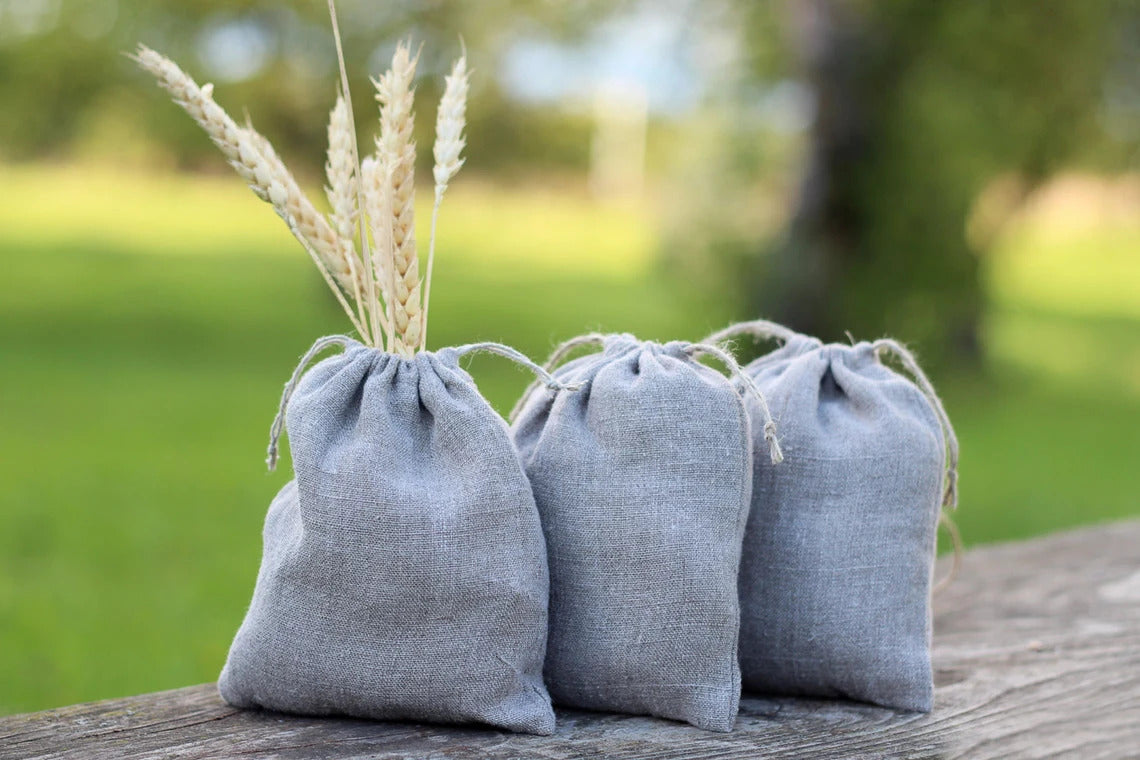 Three small natural linen gray linen drawstring pouches sit on a weathered wooden surface outdoors, with dried wheat stalks tucked into the front pouch.  