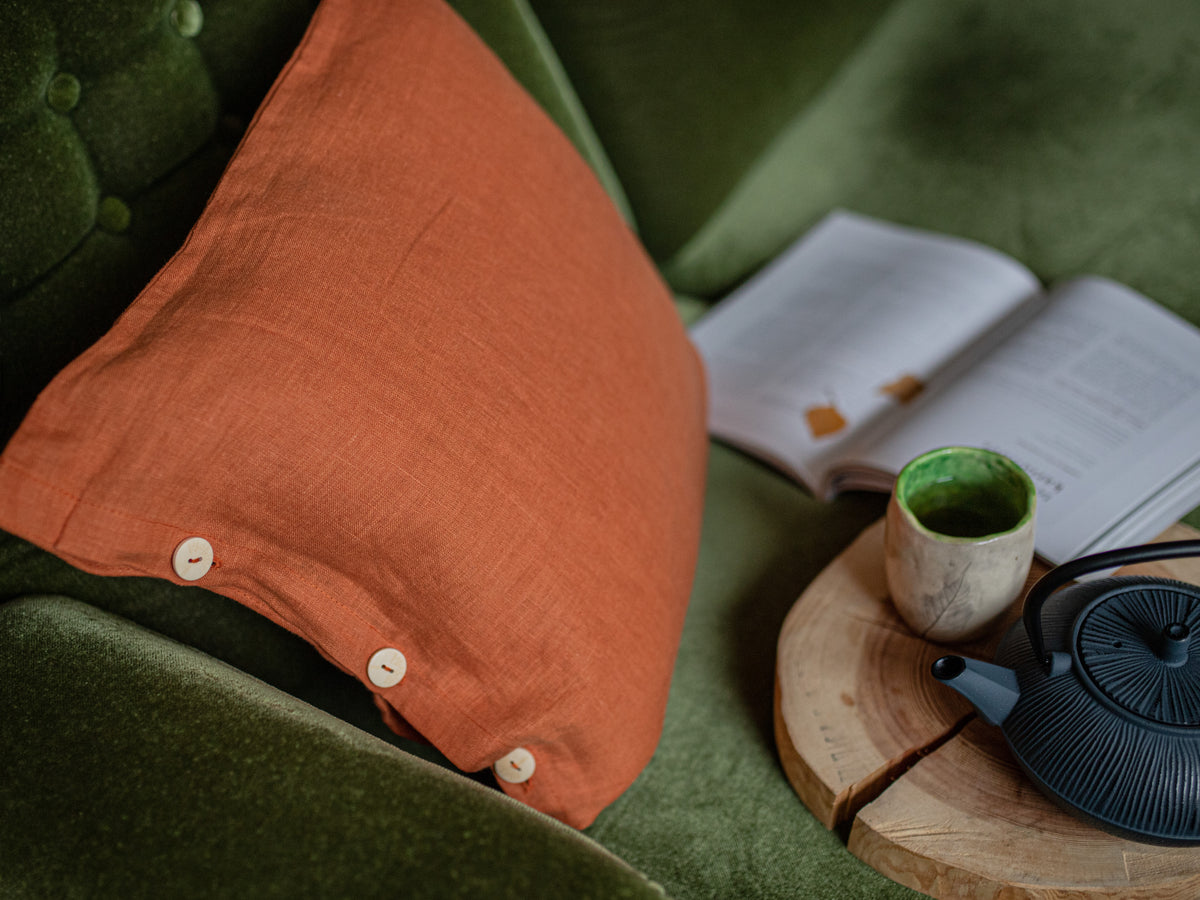 Orange pillow on a green couch with a teacup and teapot on a wooden coaster.