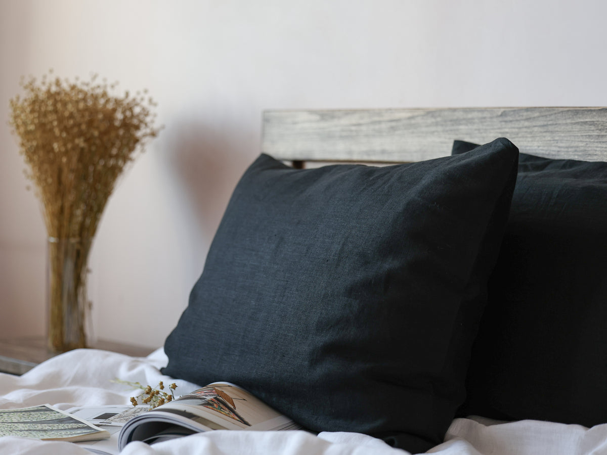 Black pillows on a bed with a magazine underneath, against a neutral wall.