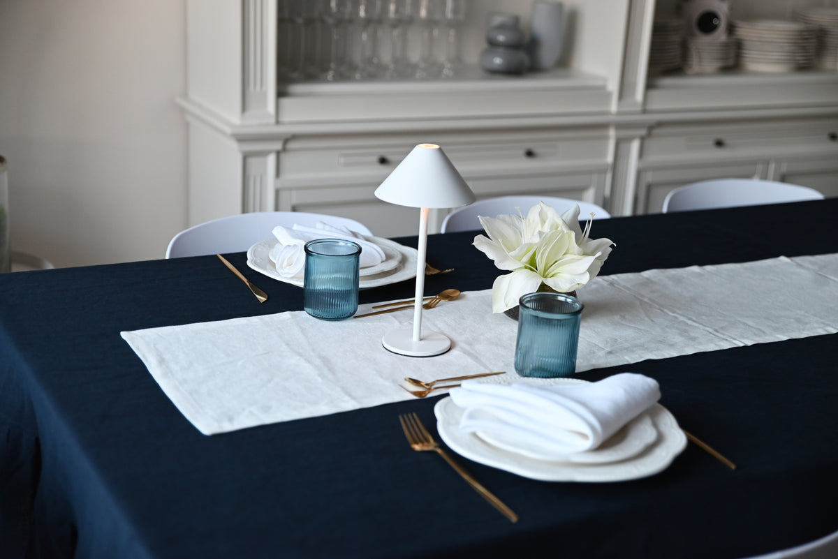 Dining table set with plates, navy blue linen tablecloth,  cutlery, and a lamp in a room.