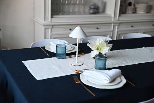 Dining table set with plates, navy blue linen tablecloth,  cutlery, and a lamp in a room.
