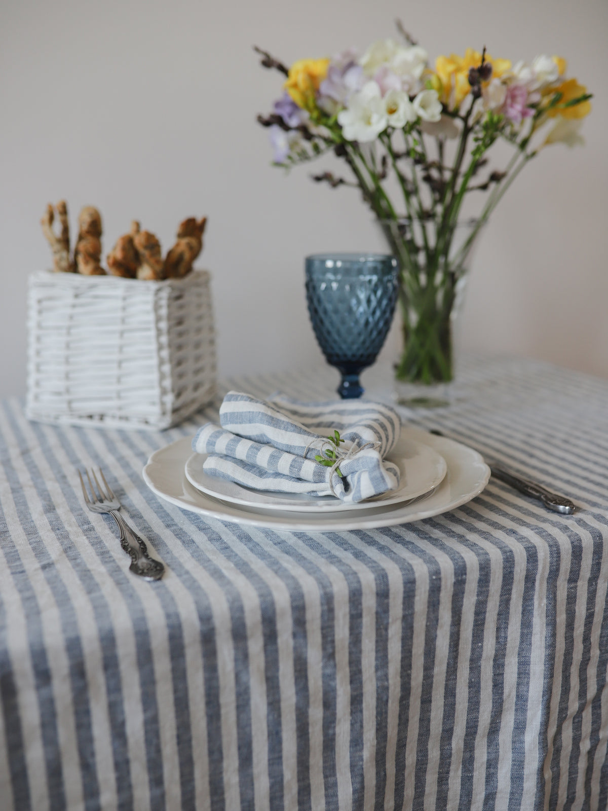 Dining table setting with striped tablecloth, plates, neutical blue linen napkins, and a vase of flowers.