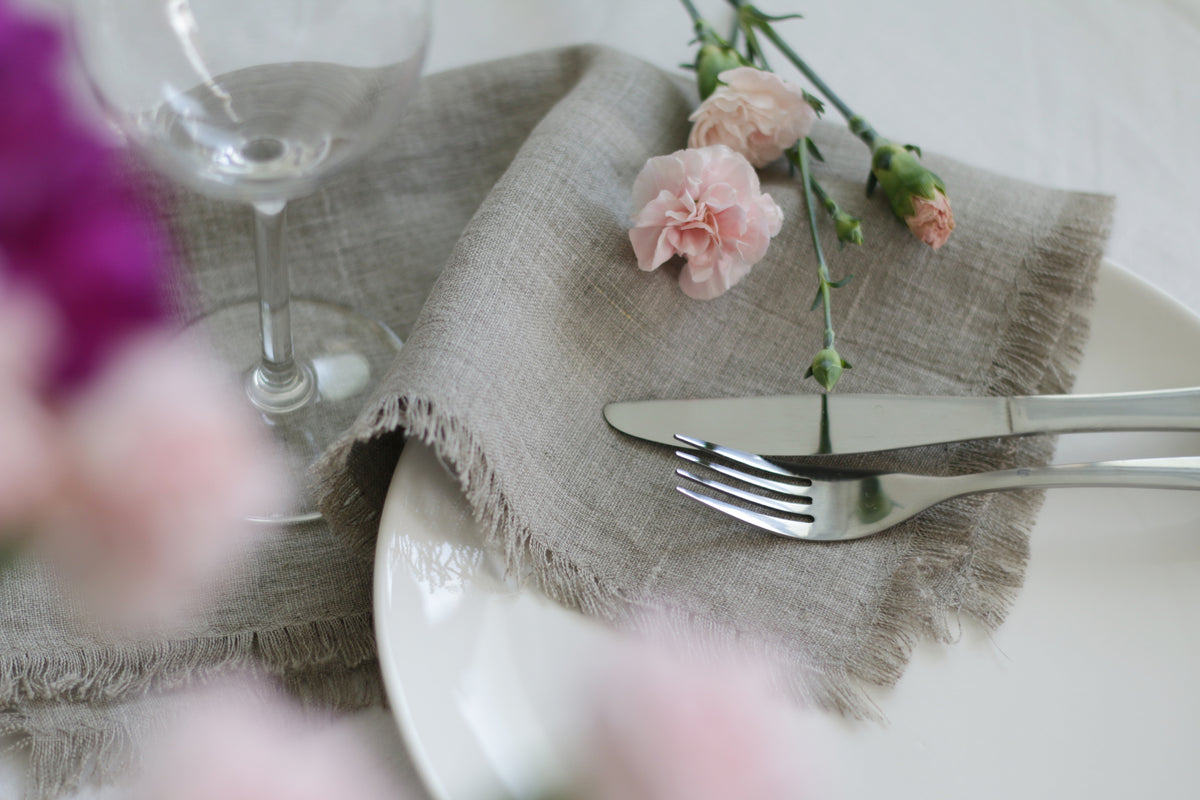 Table setting with a natural linen napkin, plate, cutlery, and flowers on a white surface.