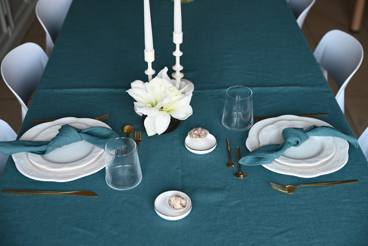Dining table set with teal linen  tablecloth, white plates, silverware, and floral centerpieces.