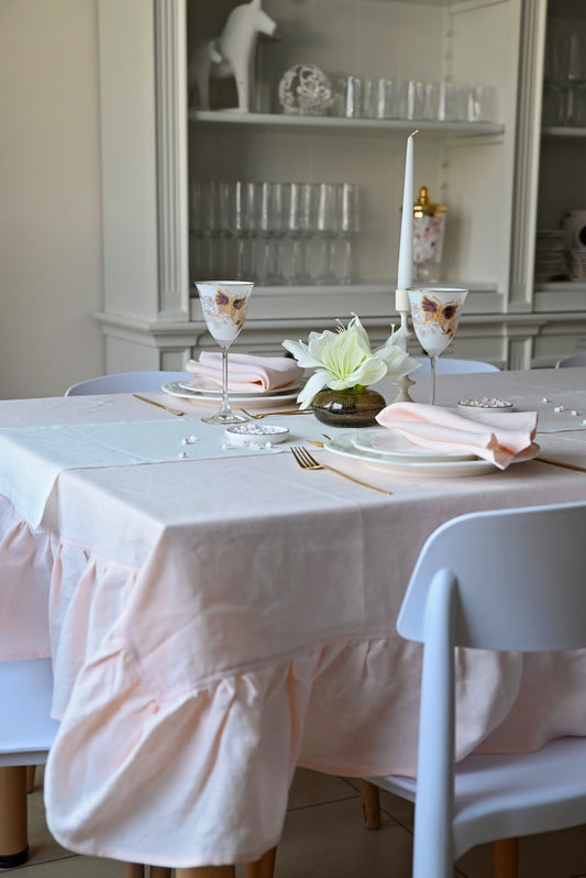 Dining table set with blush pink linen tablecloth, plates, glasses, and flowers in a room with shelves in the background.