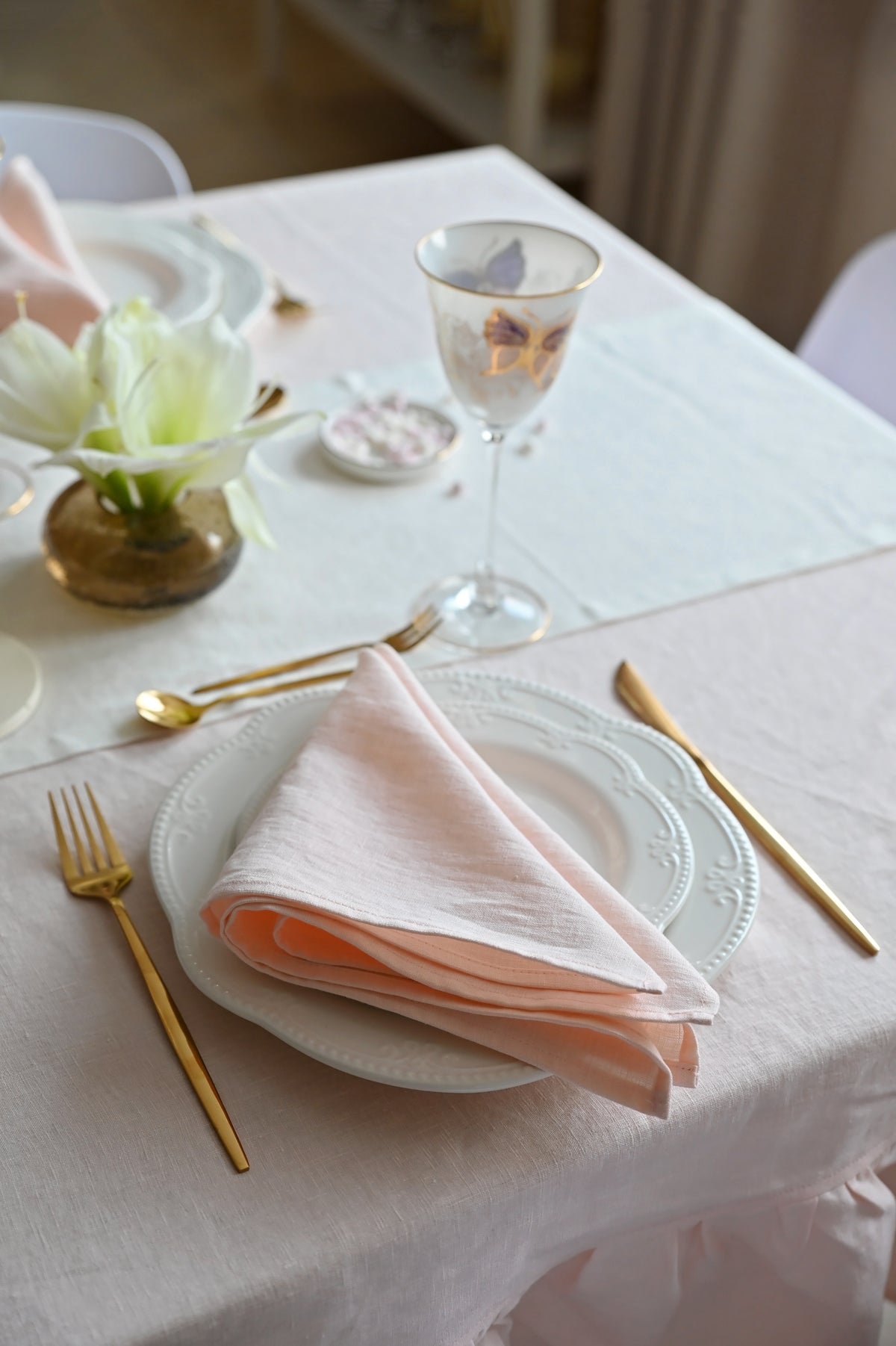 Elegant table setting with blush pink tablecloth, gold cutlery, and floral decorations.