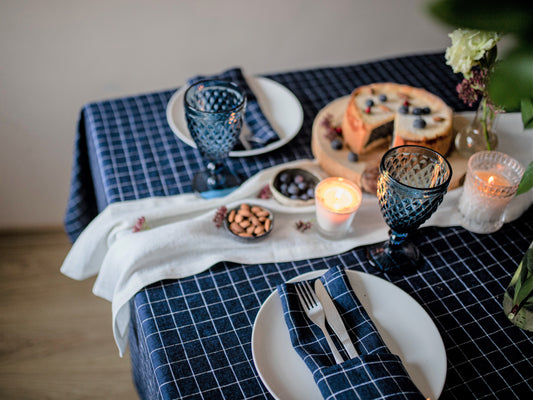 Dining table set with blue checkered linen tablecloth, plates, cutlery, candles, and a small cake.