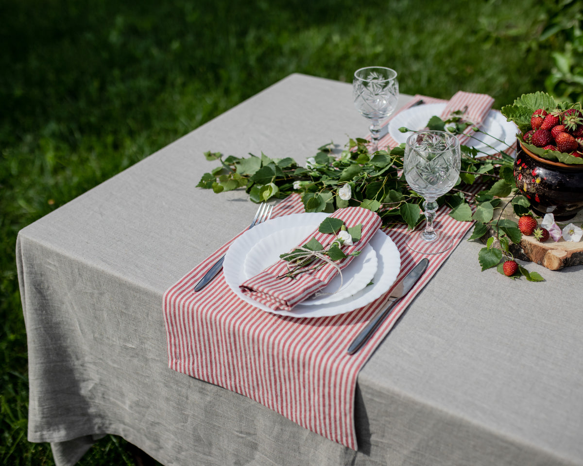 Table setting with red and white striped napkins, greenery, and glasses on a gray tablecloth outdoors.