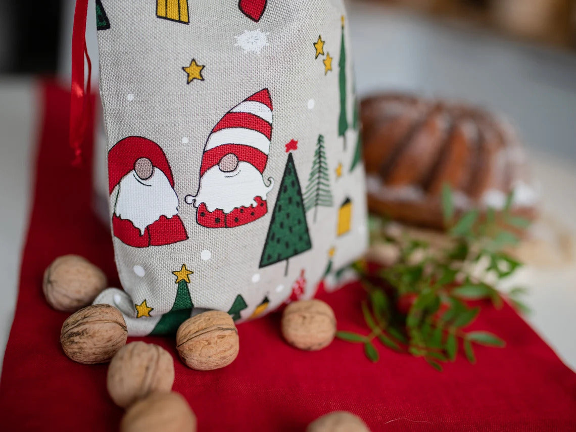 A linen fabric gift bag with a holiday gnome and Christmas tree print, styled on a red table runner with walnuts and greenery.
