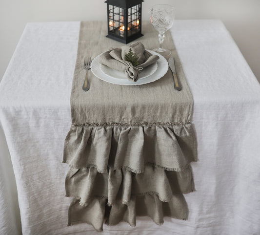 A natural linen  table runner with tiered ruffles and a delicate embroidered band, styled on a white tablecloth with a place setting, a black lantern, and a wine glass.