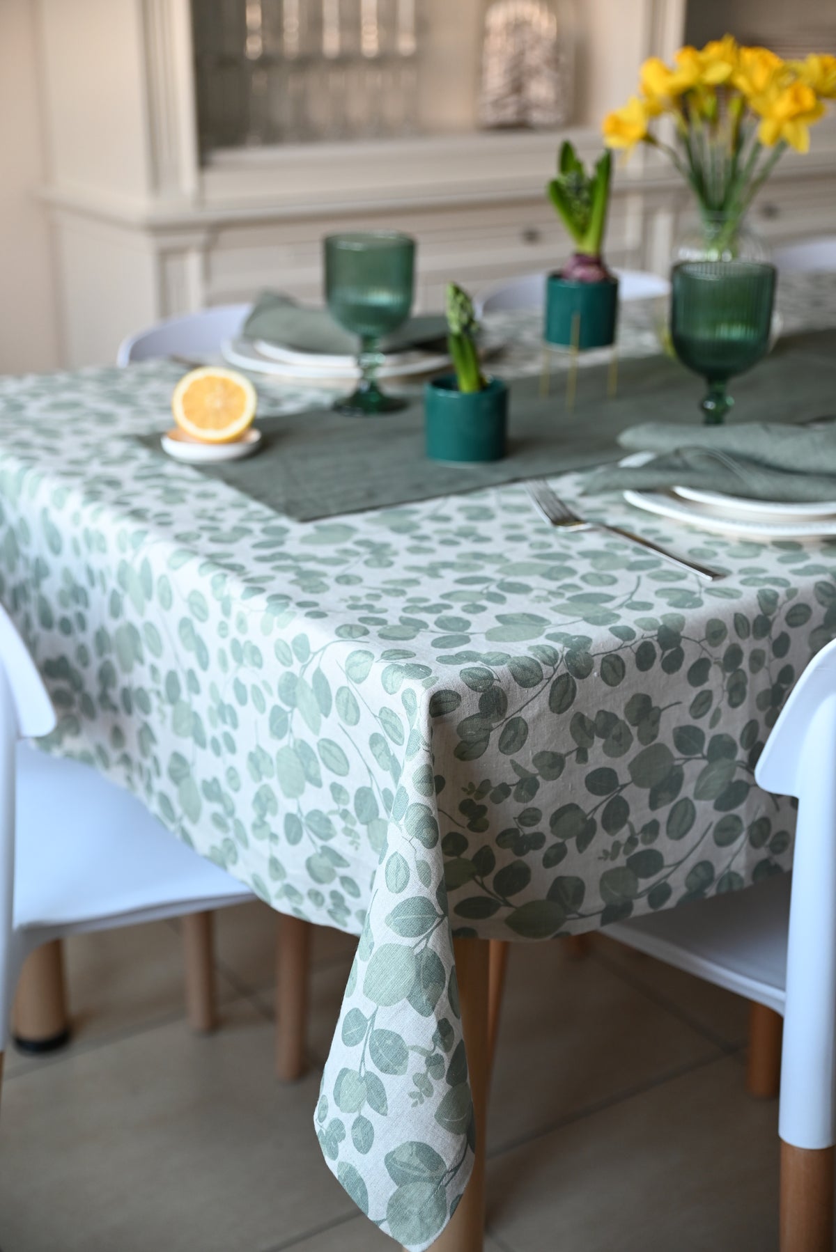 Table with a green floral linen  tablecloth, green bottles, and yellow flowers in a kitchen setting.