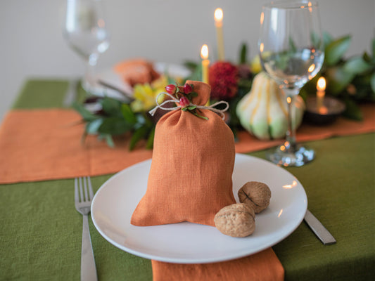 An orange linen drawstring gift bag with a small sprig of greenery and red berries, styled on a white plate with walnuts on a decorated autumn table setting.