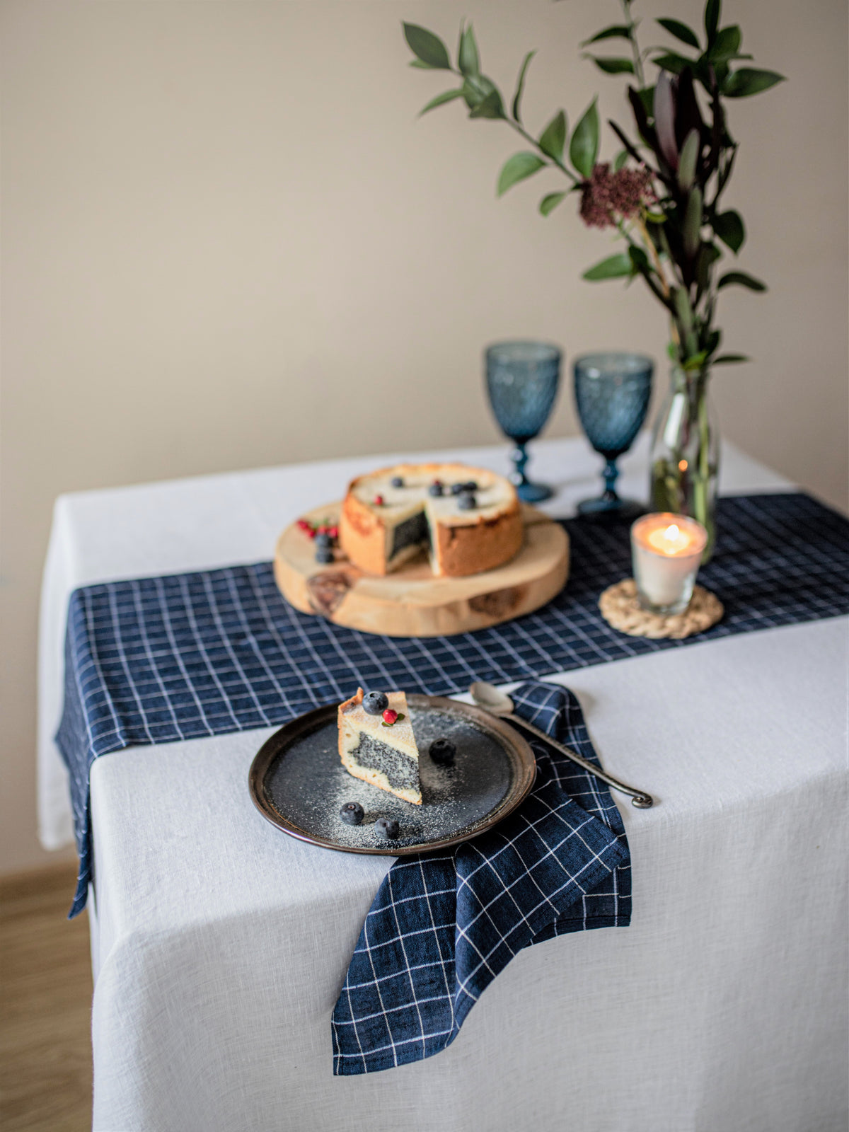 Table setting with a slice of cake on a plate, a loaf of bread, blue glasses, and a candle on a white tablecloth with a blue checkered napkin.
