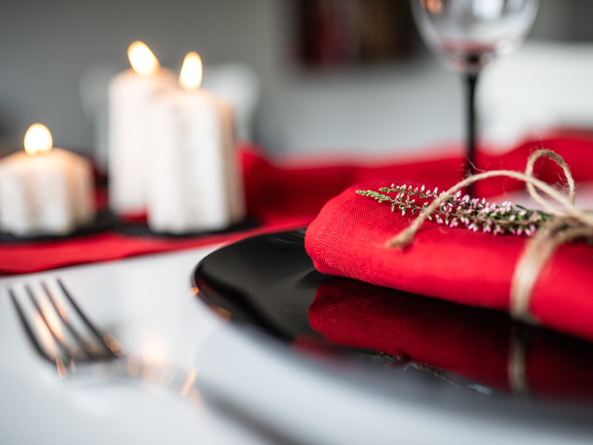 Dining table setting with red linen napkin, black plate, and candles in the background.