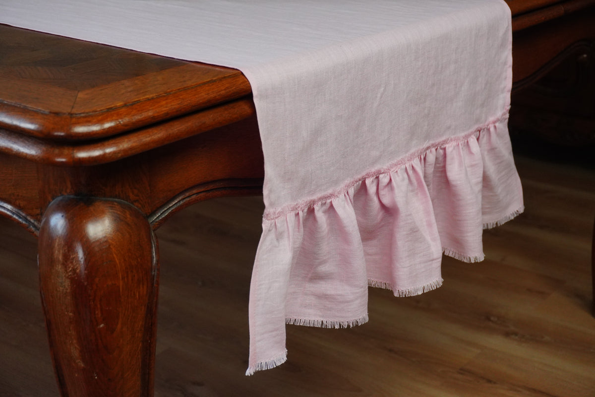 A light pink table runner with a ruffled hem and fringed edges draped over a dark wood dining table on a wooden floor.