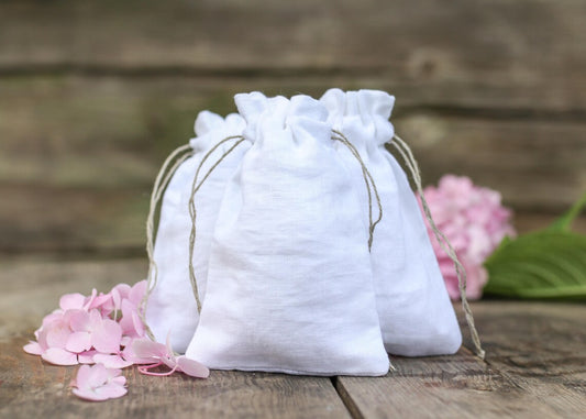 Three small white linen drawstring pouches arranged on a rustic wooden surface with pink flower petals  