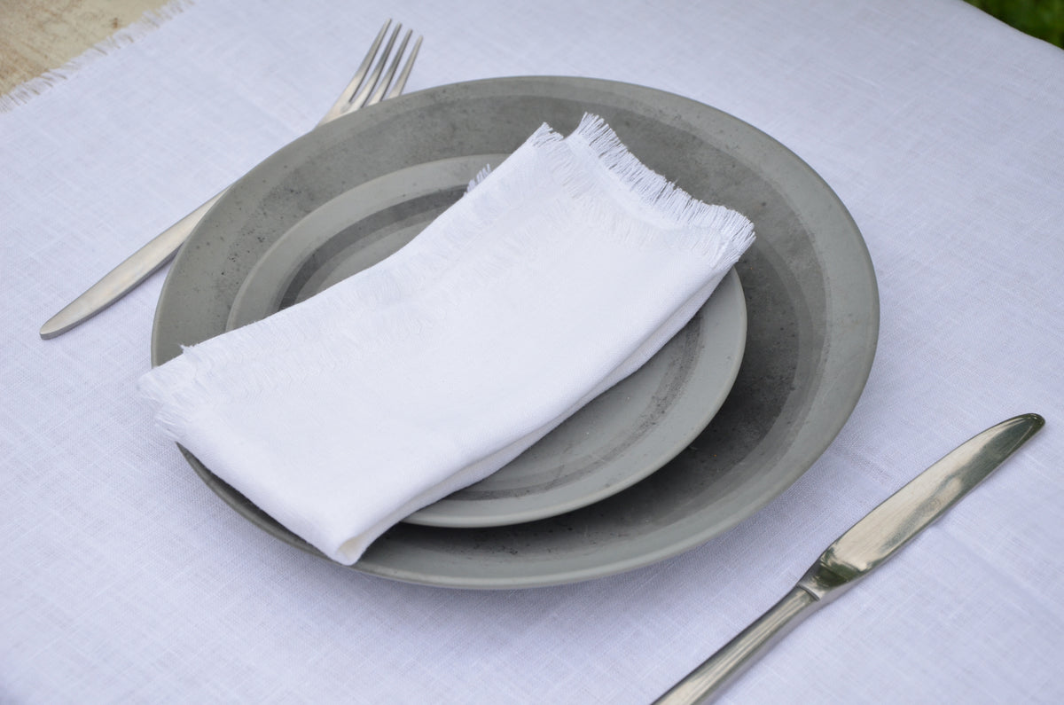 Set of gray plates with a white  fringed linen napkin, silver knife, and fork on a white tablecloth.