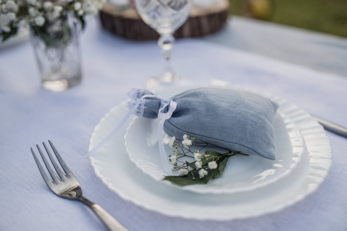 A dusty blue  linen fabric bag with a white ribbon, styled on a white plate with baby's breath flowers on a lavender tablecloth.
