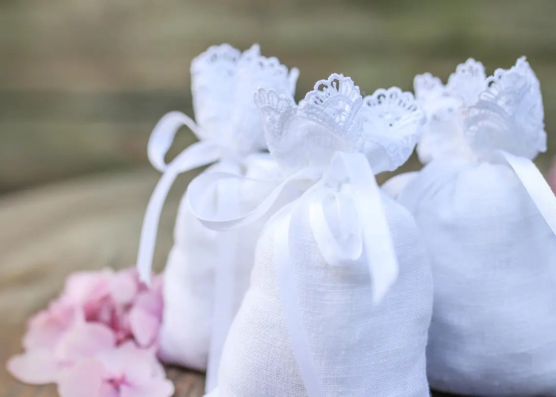 Three small white linen  pouches with lace trim and white ribbons, arranged on a wooden surface with soft pink flowers 