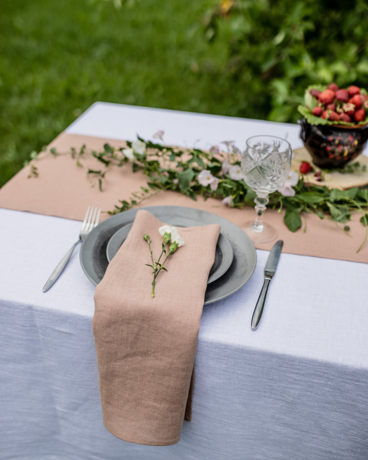 Table setting with beige napkin, gray plates, and greenery on a white tablecloth outdoors.