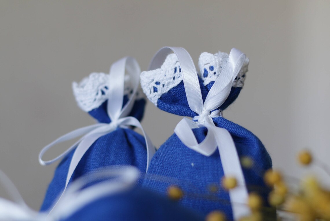 Two small blue linen pouches tied with white satin ribbons and topped with white lace trim, photographed against a soft neutral background.