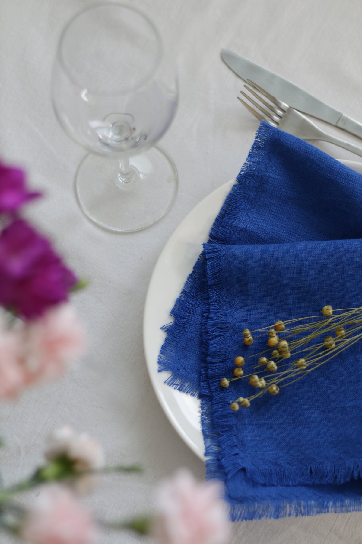 Blue fringed  linen  napkin on a white plate with a glass and flowers in the background