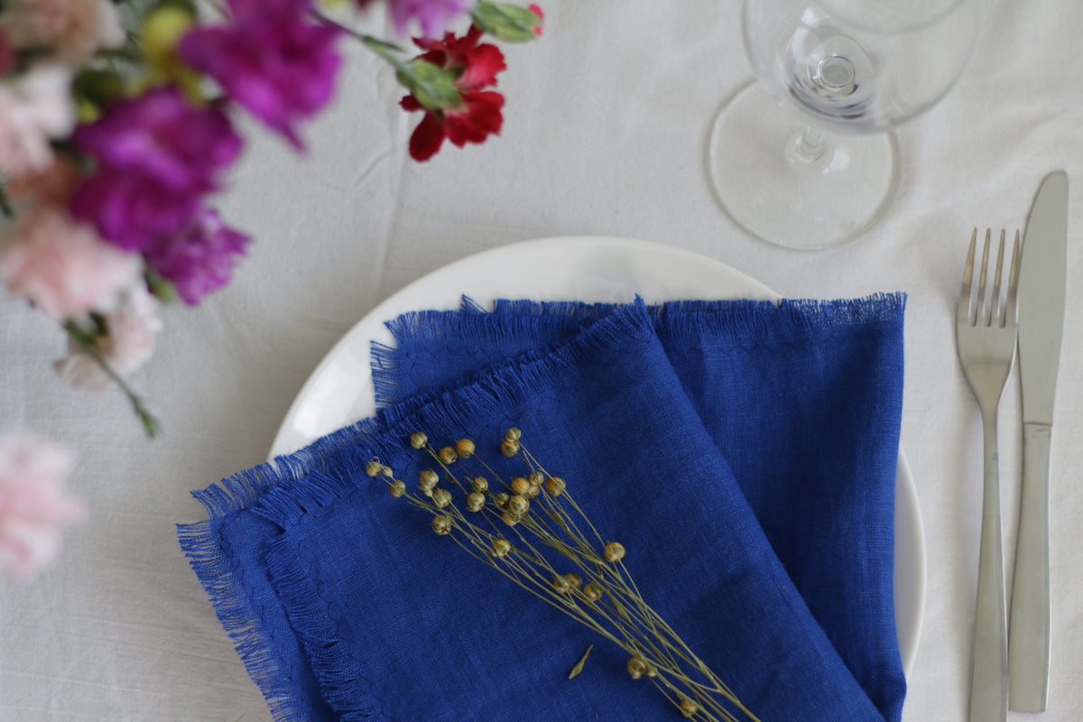 Blue linen napkin on a white plate with flowers and cutlery in the background